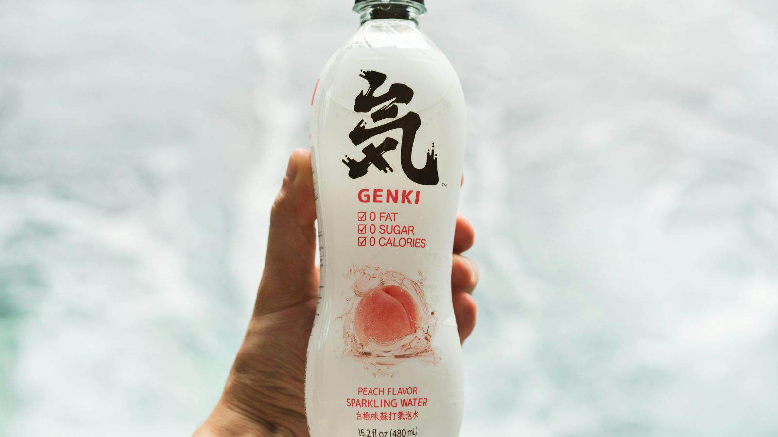 A hand holds a clear plastic bottle of Genki peach flavor sparkling water. The label shows 0 fat, 0 sugar, and 0 calories, with a peach image and large black Kanji character above the brand name.