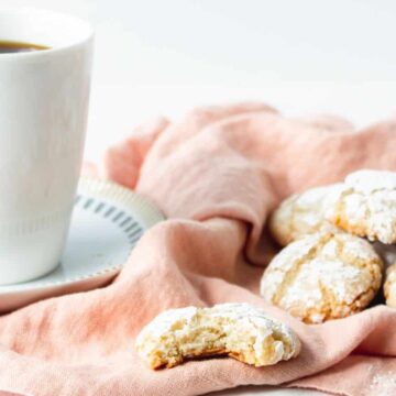 A white coffee cup on a saucer sits next to several powdered sugar cookies on a pink cloth; one cookie has a bite taken out of it.