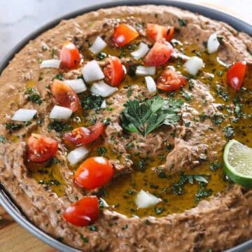 A bowl of refried beans topped with chopped tomatoes, diced onions, fresh parsley, and a lime wedge sits on a wooden board. Tortilla pieces and lime slices are visible nearby.