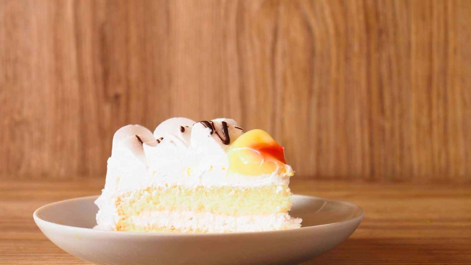 A slice of layered sponge cake with white frosting, chocolate drizzle, and a yellow fruit topping sits on a white plate against a wooden background.