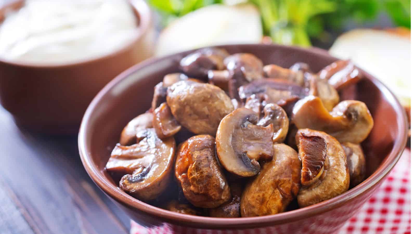 A brown bowl filled with cooked, sliced mushrooms sits on a red and white checkered cloth, with blurred greens and a bowl of white sauce in the background.