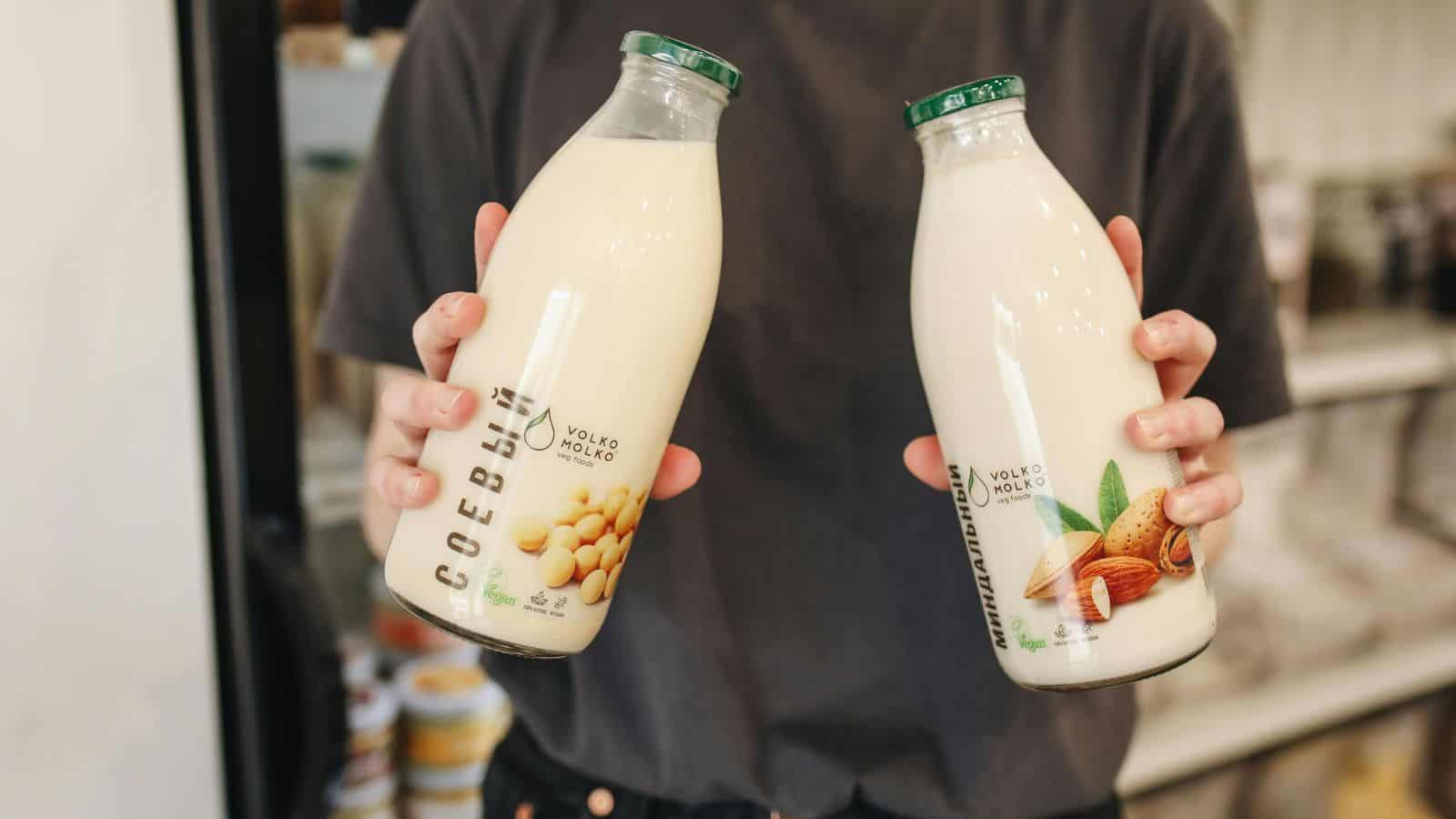 A person in a dark t-shirt holds two glass bottles of plant-based milk; the left bottle shows soybeans and the right bottle shows almonds, indicating soy milk and almond milk, respectively.