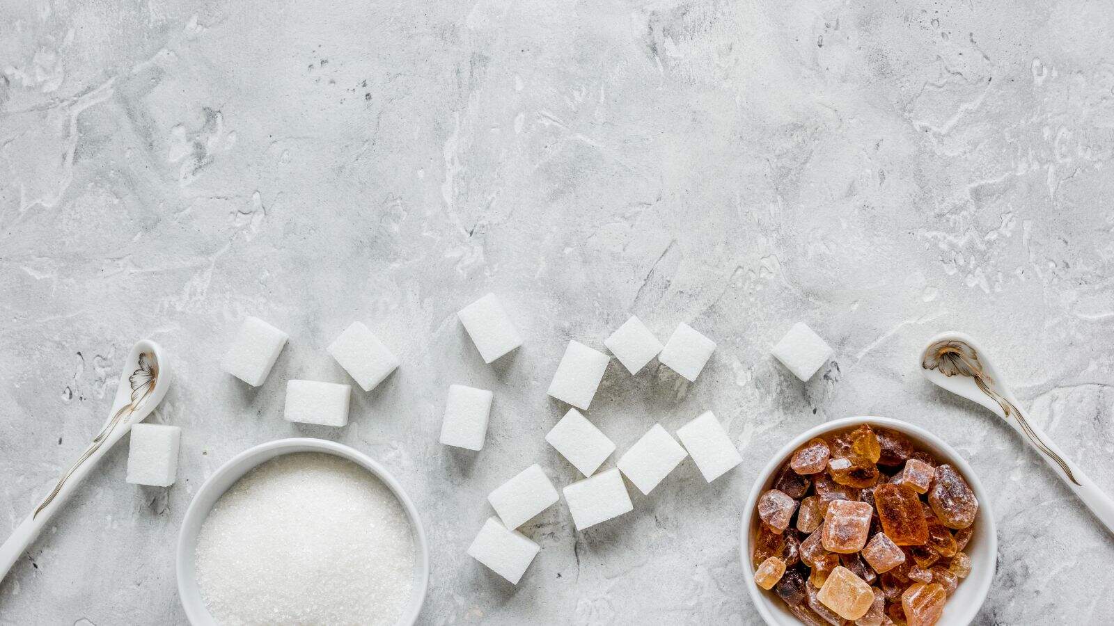 Two bowls hold white granulated sugar and brown sugar crystals, each with a spoon. Between them, white sugar cubes are scattered on a light grey textured surface.