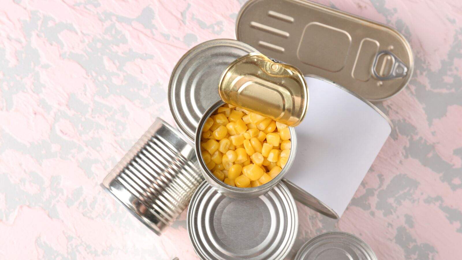 An open tin can filled with yellow corn kernels is surrounded by four unopened tin cans on a light pink and gray textured surface. One can has a blank white label.