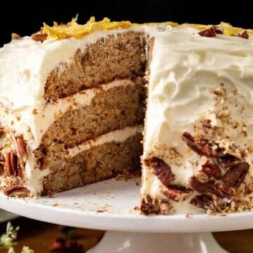 A three-layer cake with creamy white frosting, decorated with chopped pecans on the sides, sits on a white cake stand. A large slice is missing, revealing the cake’s moist, textured interior.