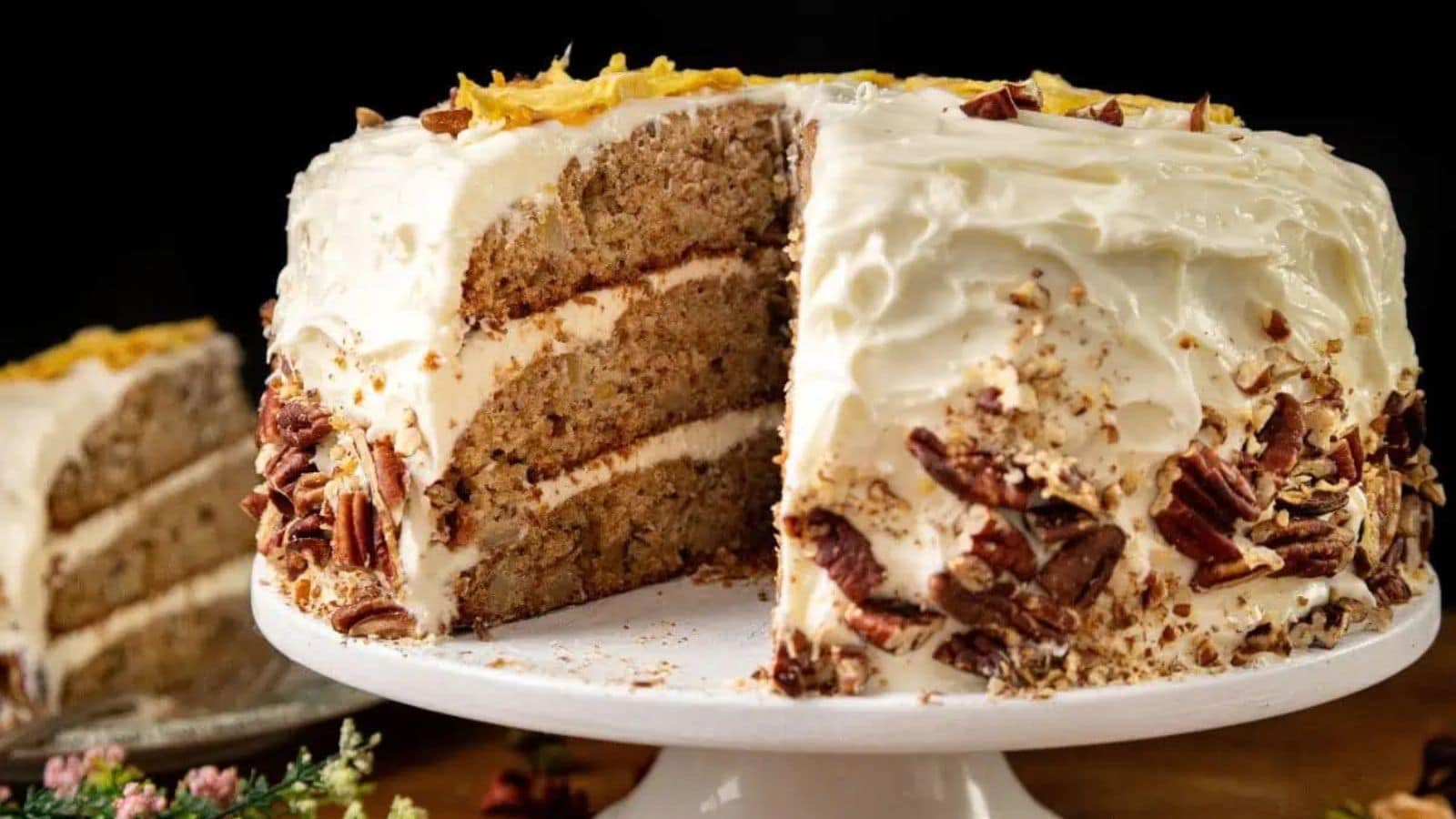 A slice of Hummingbird Cake with cream cheese frosting and pineapple chunks on top, placed on a decorative green and white plate. The Southern cake appears moist with visible nuts, and a fork is resting at the base.