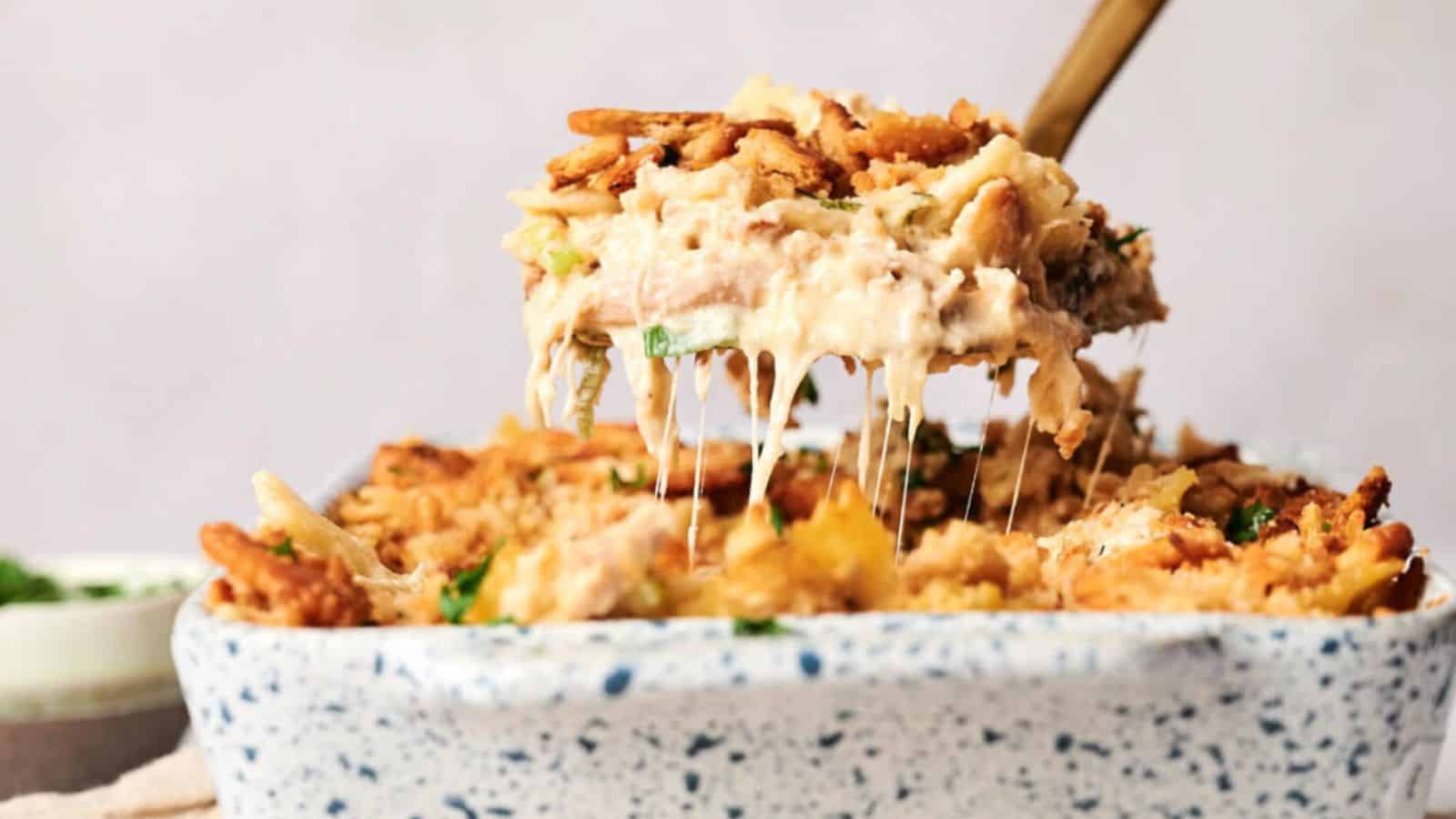 A wooden spoon lifts a cheesy, creamy casserole topped with crispy fried onions from a speckled baking dish. The casserole has visible layers and melted cheese stretching from the dish.