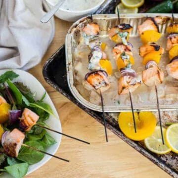 Grilled salmon, yellow bell pepper, and red onion skewers on a baking tray, with lemon slices and jalapeños nearby. A plate with salad greens and additional skewers sits to the side.