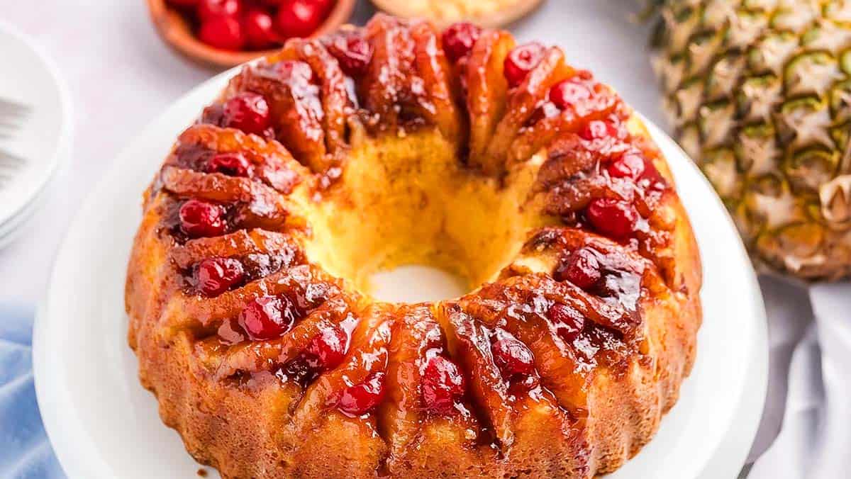 A round pineapple upside-down cake topped with red cherries, displayed on a white plate. Pineapple and cherries in the background.