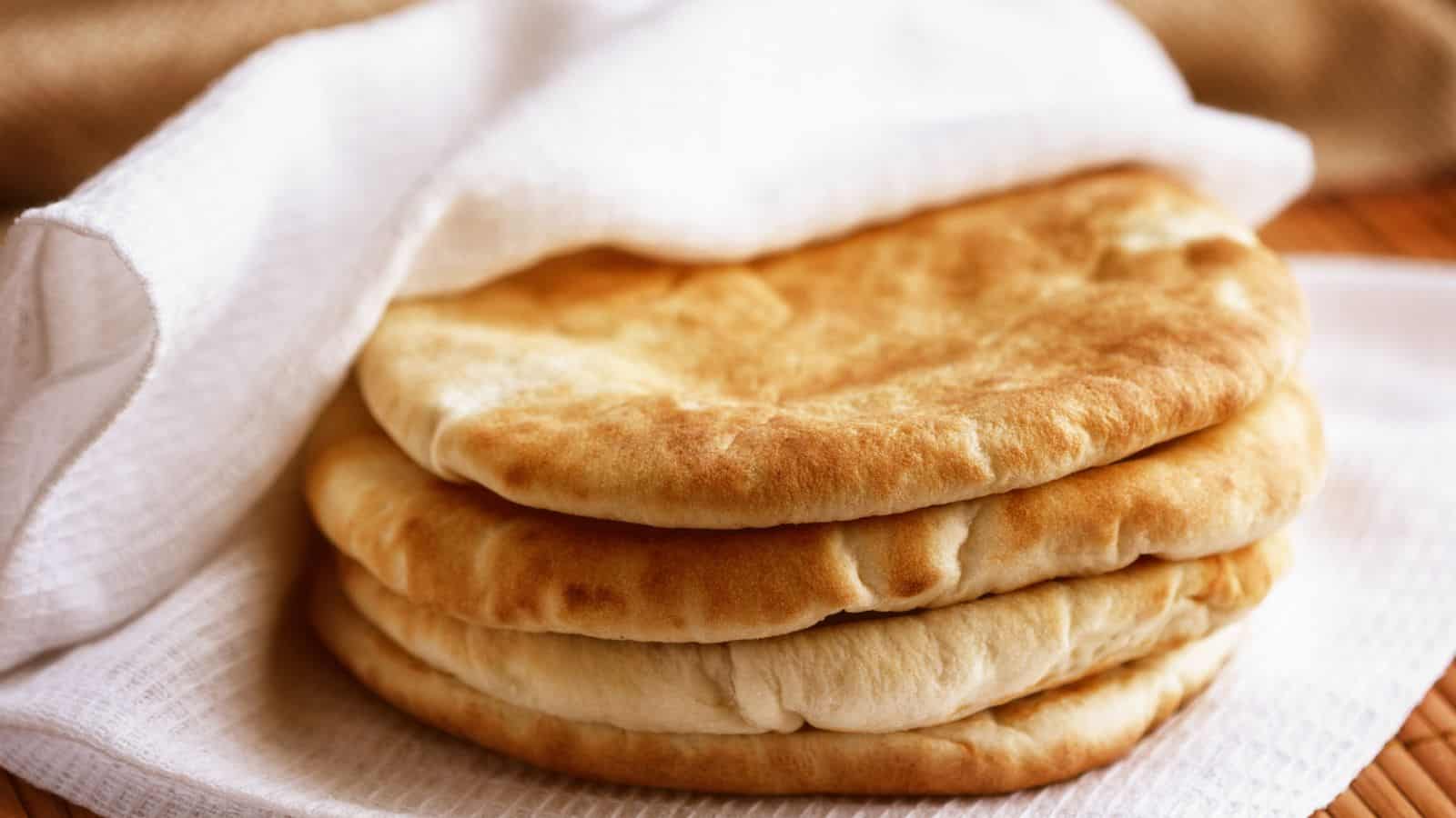 A stack of three pita breads sits partially covered by a white cloth on a wooden surface.