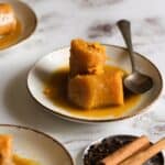 Two pieces of cooked plantain in syrup, reminiscent of Plantains in Coconut Milk, are served on a white plate with a spoon. Cinnamon sticks and cloves sit nearby on the light, marbled surface.