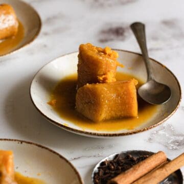 Two pieces of cooked plantain in syrup, reminiscent of Plantains in Coconut Milk, are served on a white plate with a spoon. Cinnamon sticks and cloves sit nearby on the light, marbled surface.