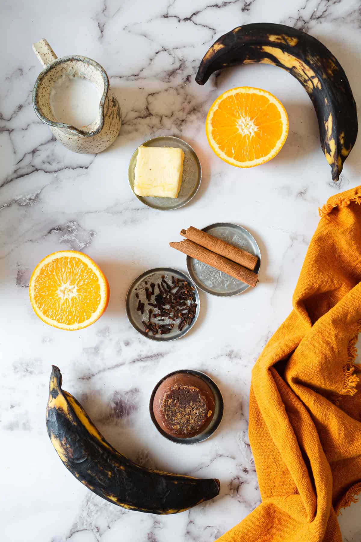 Overripe bananas, orange halves, a small jug of milk, a pat of butter, cinnamon sticks, cloves, ground spice in a small bowl, and an orange cloth evoke the comforting flavors found in Plantains in Coconut Milk.