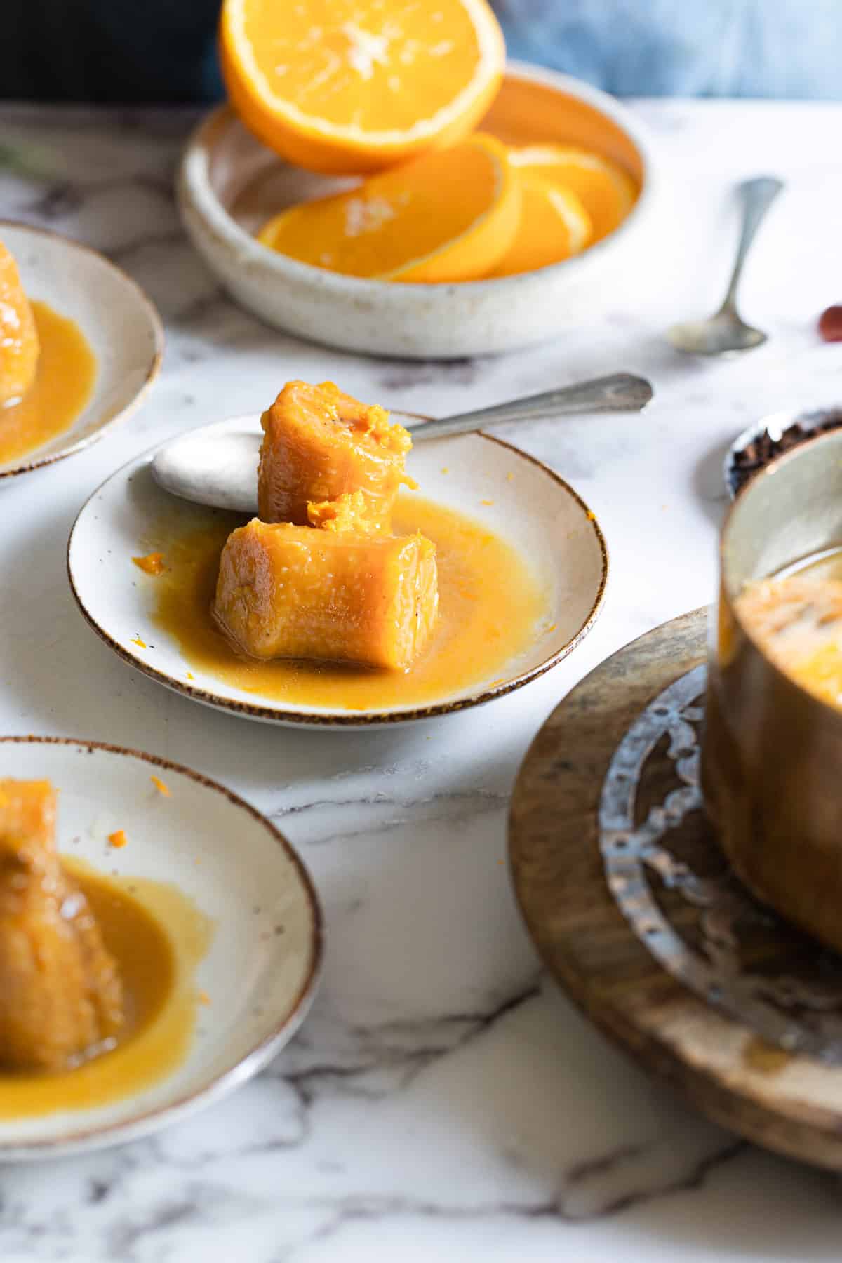 A small plate with pieces of steamed cassava covered in orange syrup sits on a marble surface next to a spoon, evoking the tropical charm of plantains in coconut milk. In the background, orange slices rest in a bowl beside a metal pot.