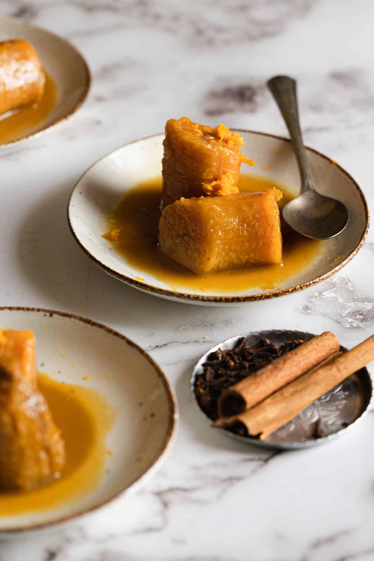 A small plate with pieces of pumpkin in syrup and a spoon, next to a dish containing cinnamon sticks and star anise, evokes the comforting flavors of Plantains in Coconut Milk, all set on a white marble surface.