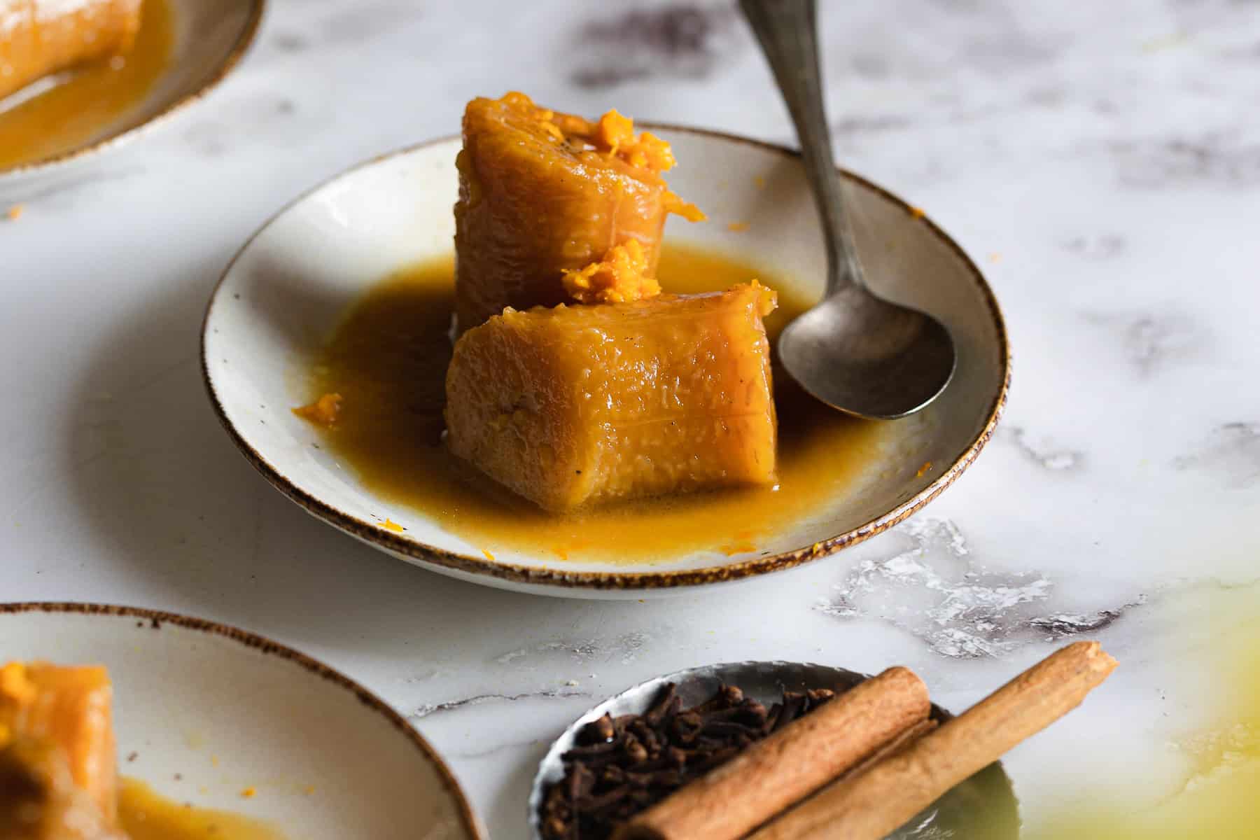 Two pieces of caramelized plantain in syrup, reminiscent of Plantains in Coconut Milk, are served on a white plate with a spoon. Nearby, a small dish with whole cloves and a cinnamon stick rests on the marble surface.