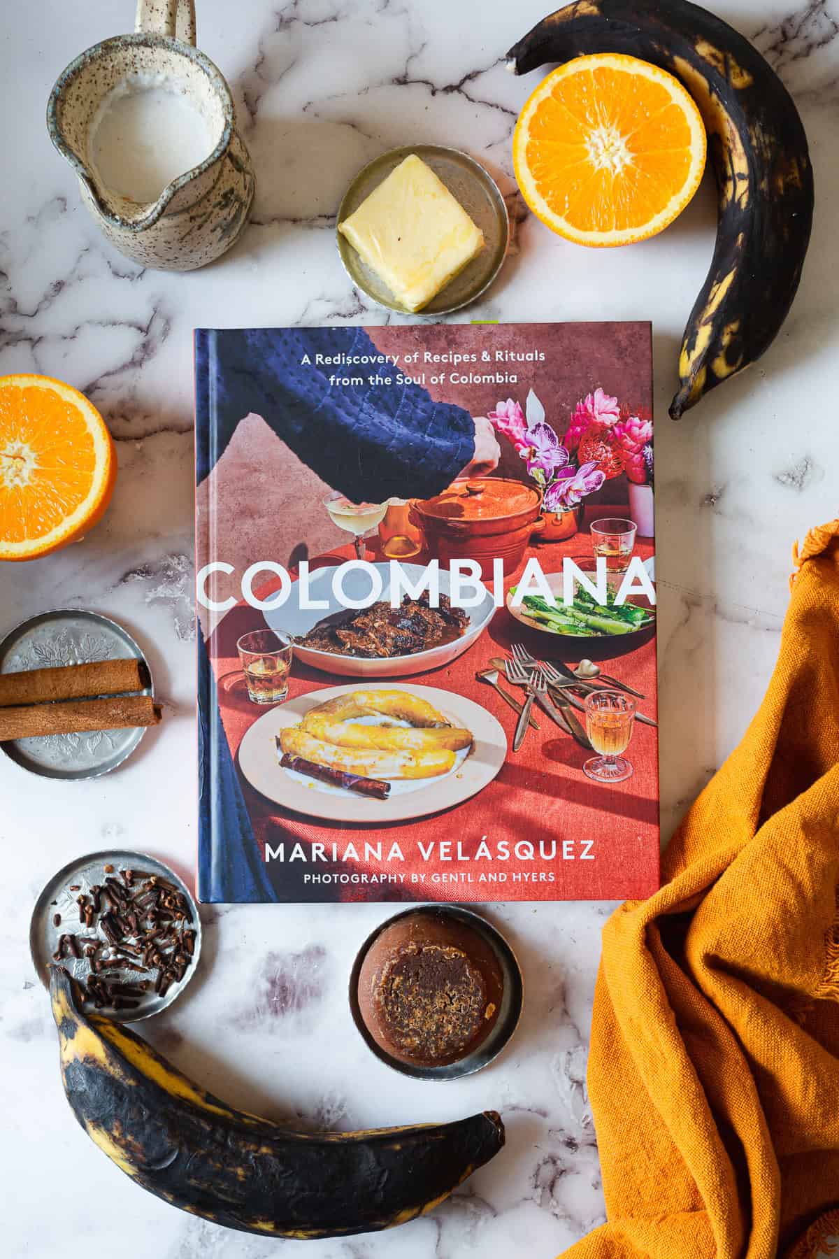 A cookbook titled "Colombiana" by Mariana Velásquez rests on a marble surface, surrounded by ripe plantains, orange slices, cheese, spices, a jar of cream, and an orange cloth—perfect inspiration for Plantains in Coconut Milk.