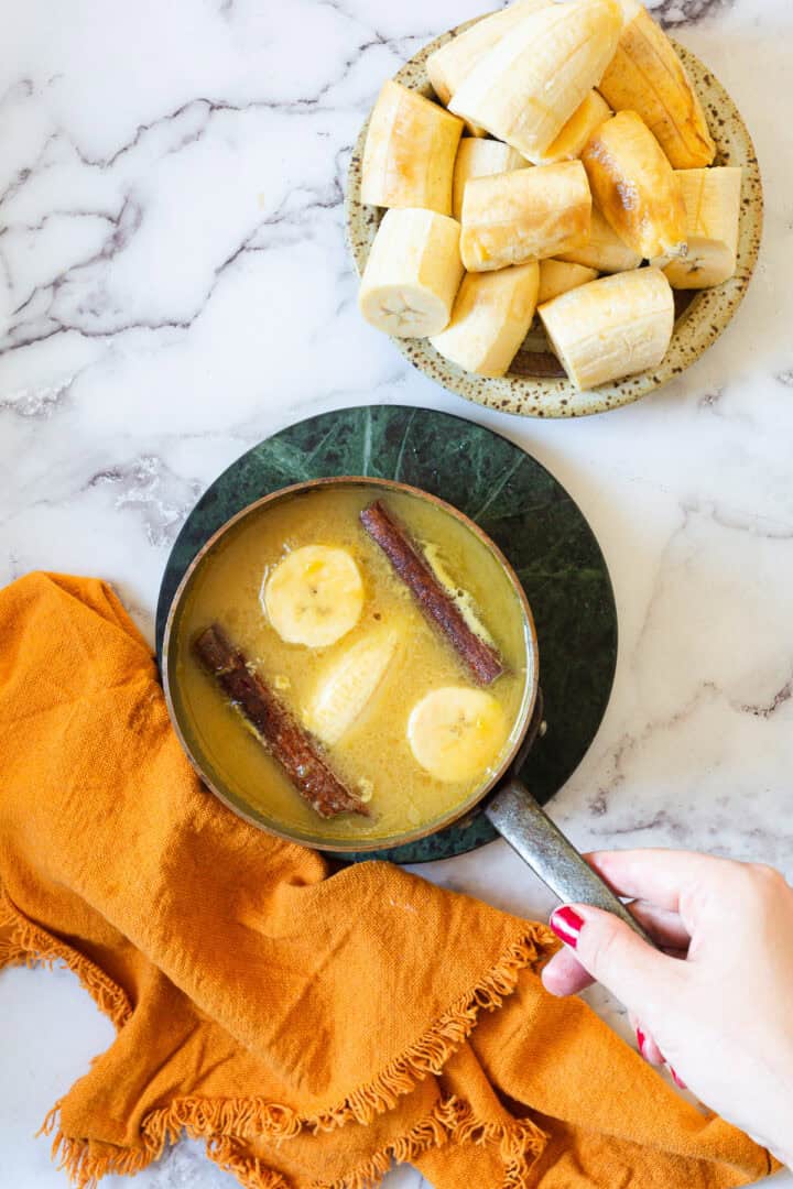 A hand holds a pot filled with banana slices, whole cinnamon sticks, and a yellow liquid—perfect for making Plantains in Coconut Milk. Nearby, an orange cloth and a plate of more sliced bananas rest on the marble surface.