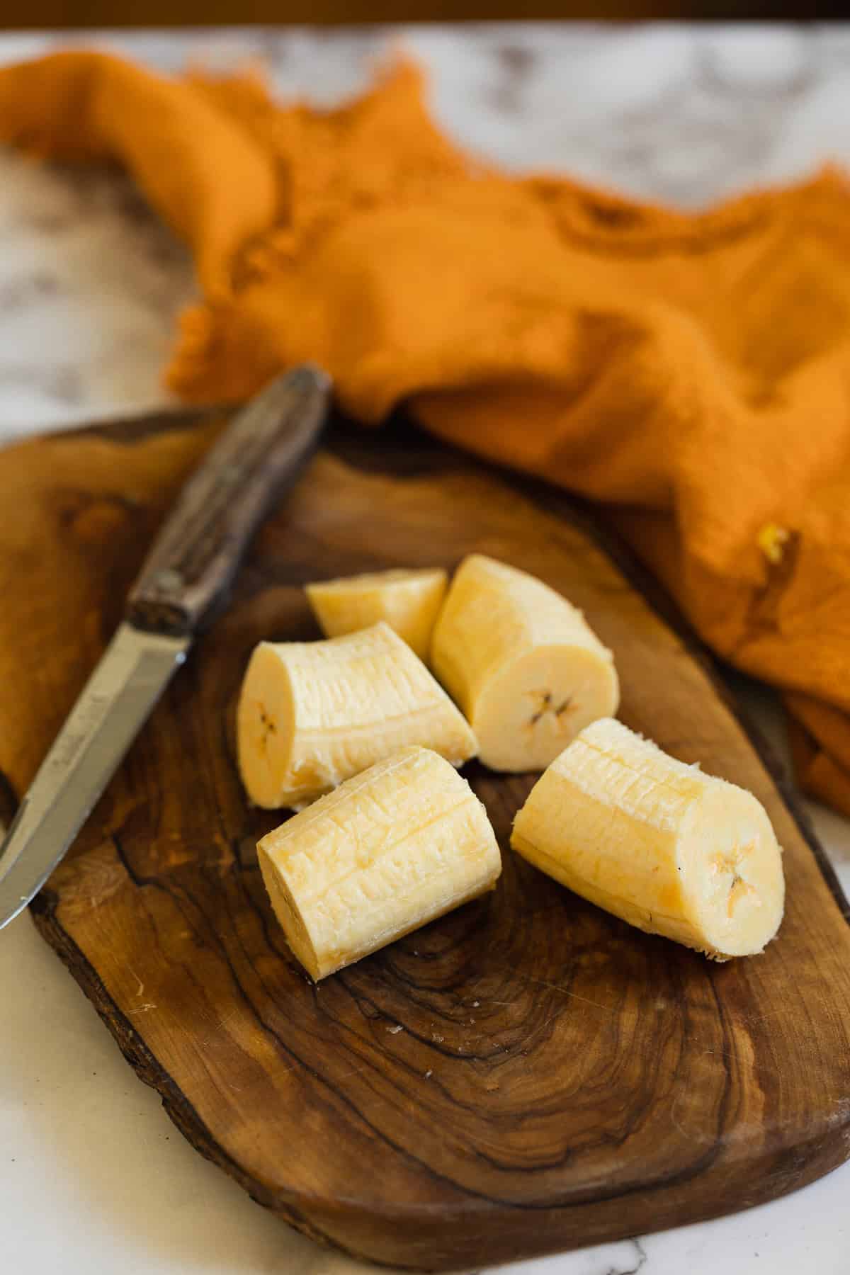 A peeled plantain cut into several chunks sits on a wooden cutting board next to a knife, ready for Plantains in Coconut Milk; an orange cloth is in the background.