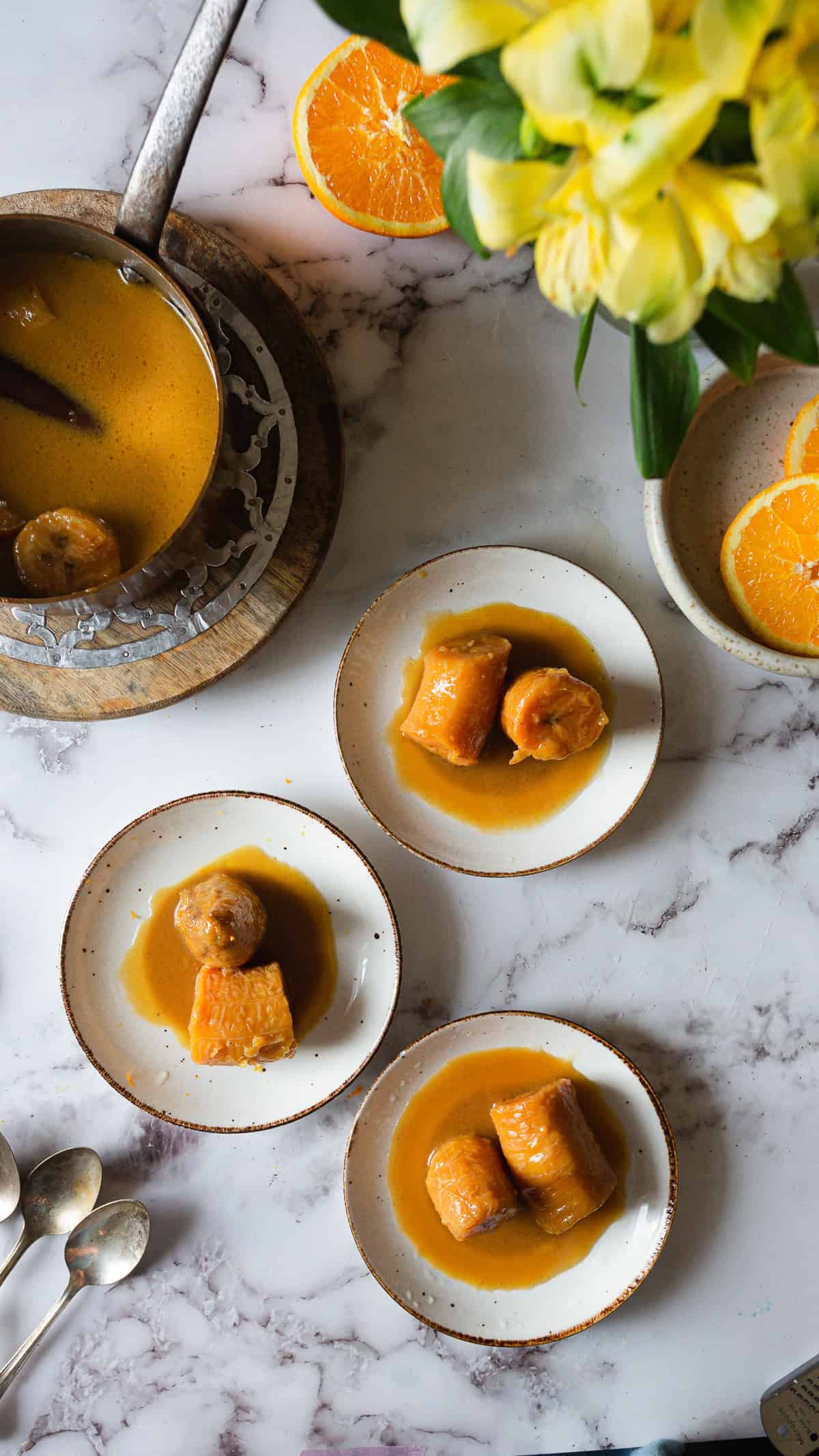Three small plates with pieces of sausage in orange sauce are arranged on a marble surface. A pot with more sauce, orange slices, yellow flowers, and two spoons create a vibrant scene reminiscent of Plantains in Coconut Milk desserts.