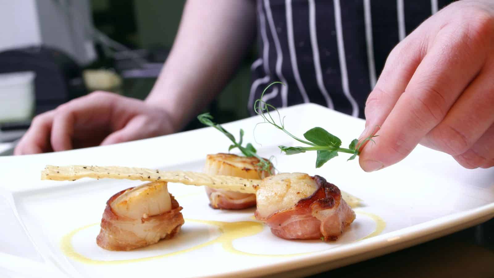 A person garnishes a plated dish of three bacon-wrapped scallops with a sprig of greens. The scallops are arranged on a white plate with sauce and a crisp bread stick. The person wears a striped apron.