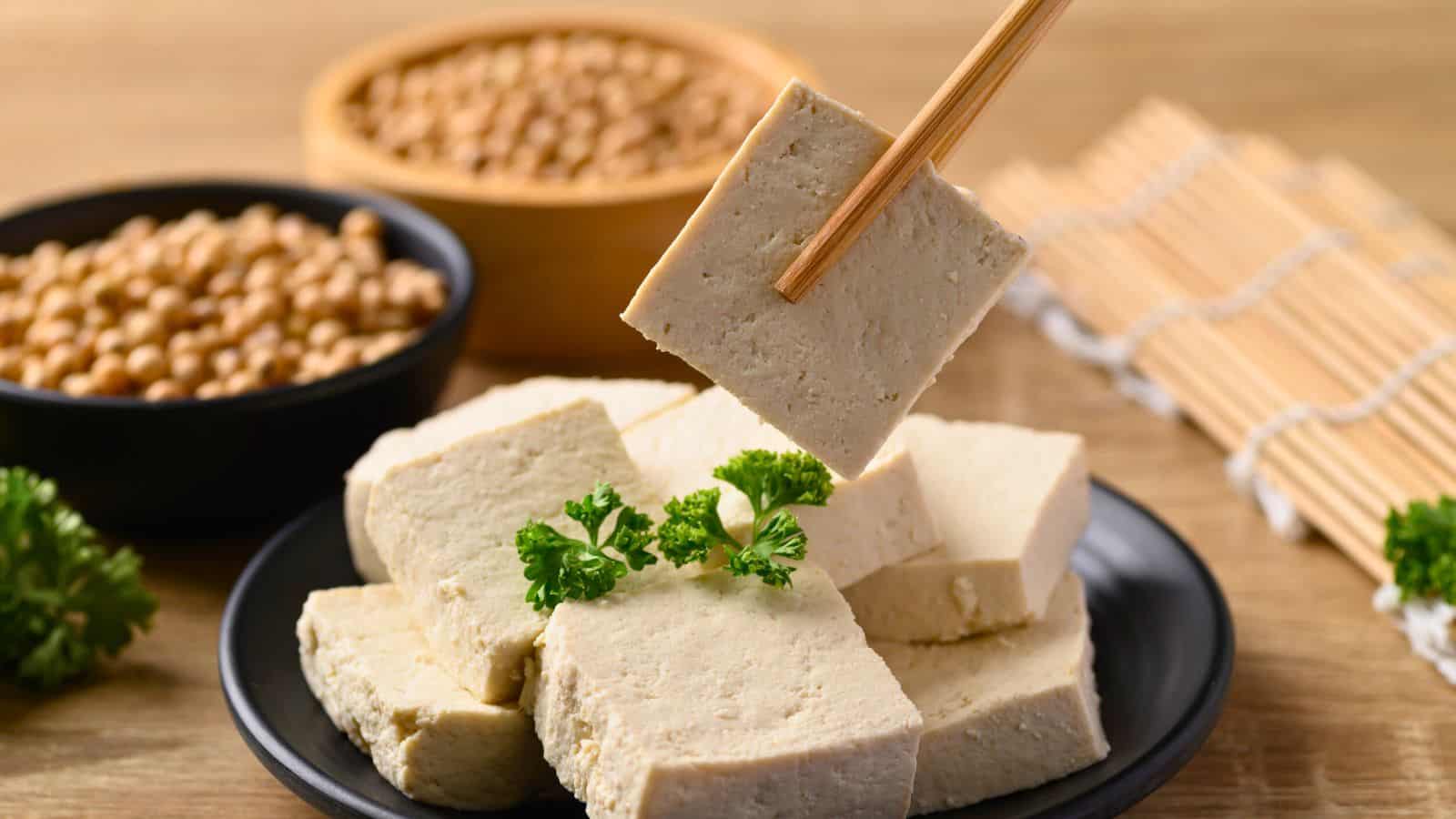 A plate of tofu blocks garnished with parsley sits on a table. One tofu block is being held by chopsticks above the plate. In the background are bowls of soybeans and a bamboo sushi rolling mat.