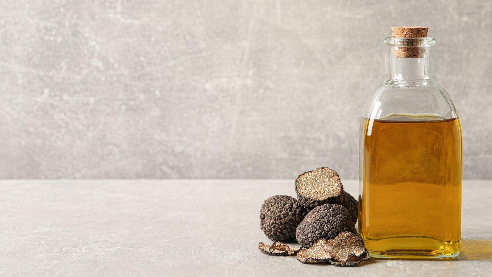 A glass bottle of golden truffle oil with a cork sits next to whole and sliced black truffles on a light gray surface against a textured gray background.