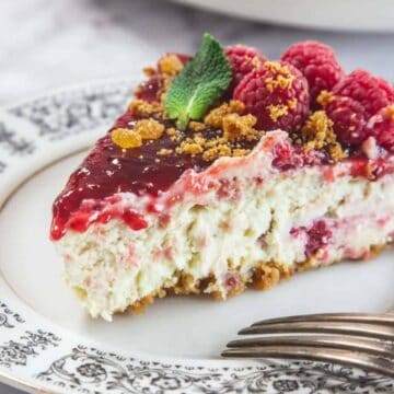 A slice of raspberry cheesecake topped with raspberries, mint leaves, and crumbled topping is placed on a decorative plate with a fork beside it.