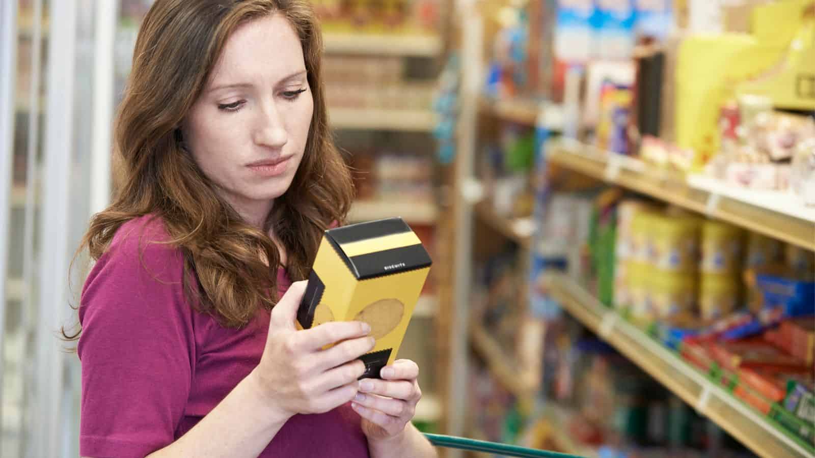 A woman with long brown hair wearing a maroon shirt stands in a grocery store aisle, holding and reading the label on a yellow and black box. Shelves with various food items are visible in the background.