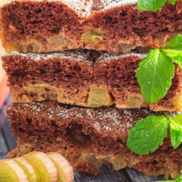 Three stacked slices of rhubarb cake are dusted with powdered sugar and garnished with fresh mint leaves, with rhubarb pieces and stalks placed nearby on a dark wooden surface.