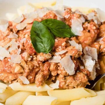 A white bowl filled with penne pasta topped with a meat and tomato sauce, garnished with grated parmesan cheese and fresh basil leaves, with a fork resting in the bowl.