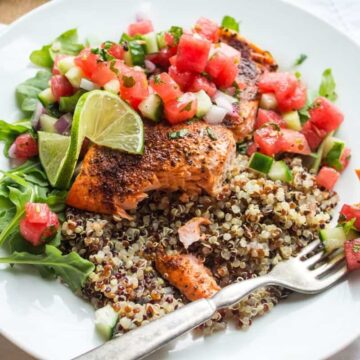 A white plate with cooked salmon fillet, quinoa, arugula, watermelon and cucumber salsa, and a lime wedge. A fork rests on the plate with a piece of salmon on its tines.