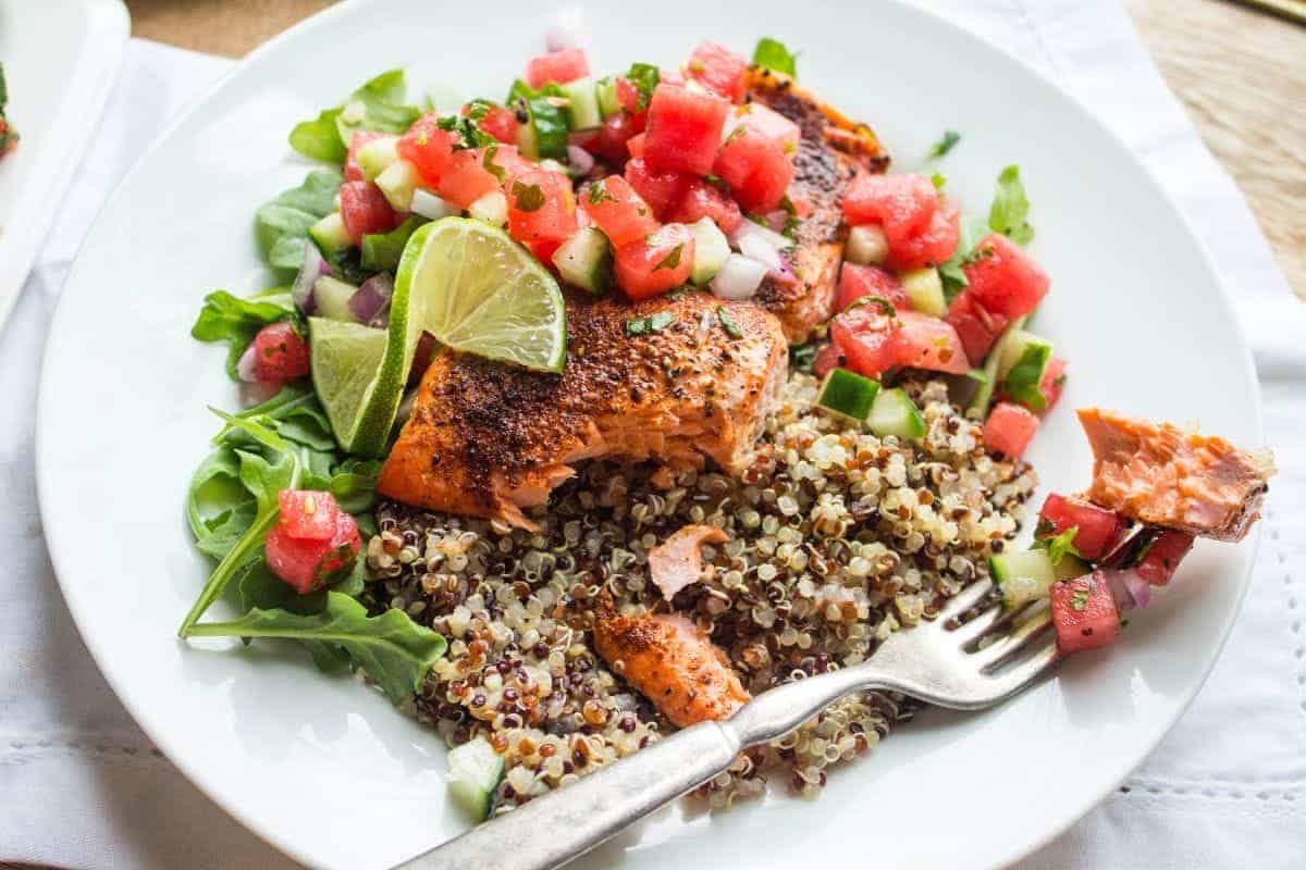 A white plate with quinoa, arugula, and roasted salmon topped with a watermelon and cucumber salsa.