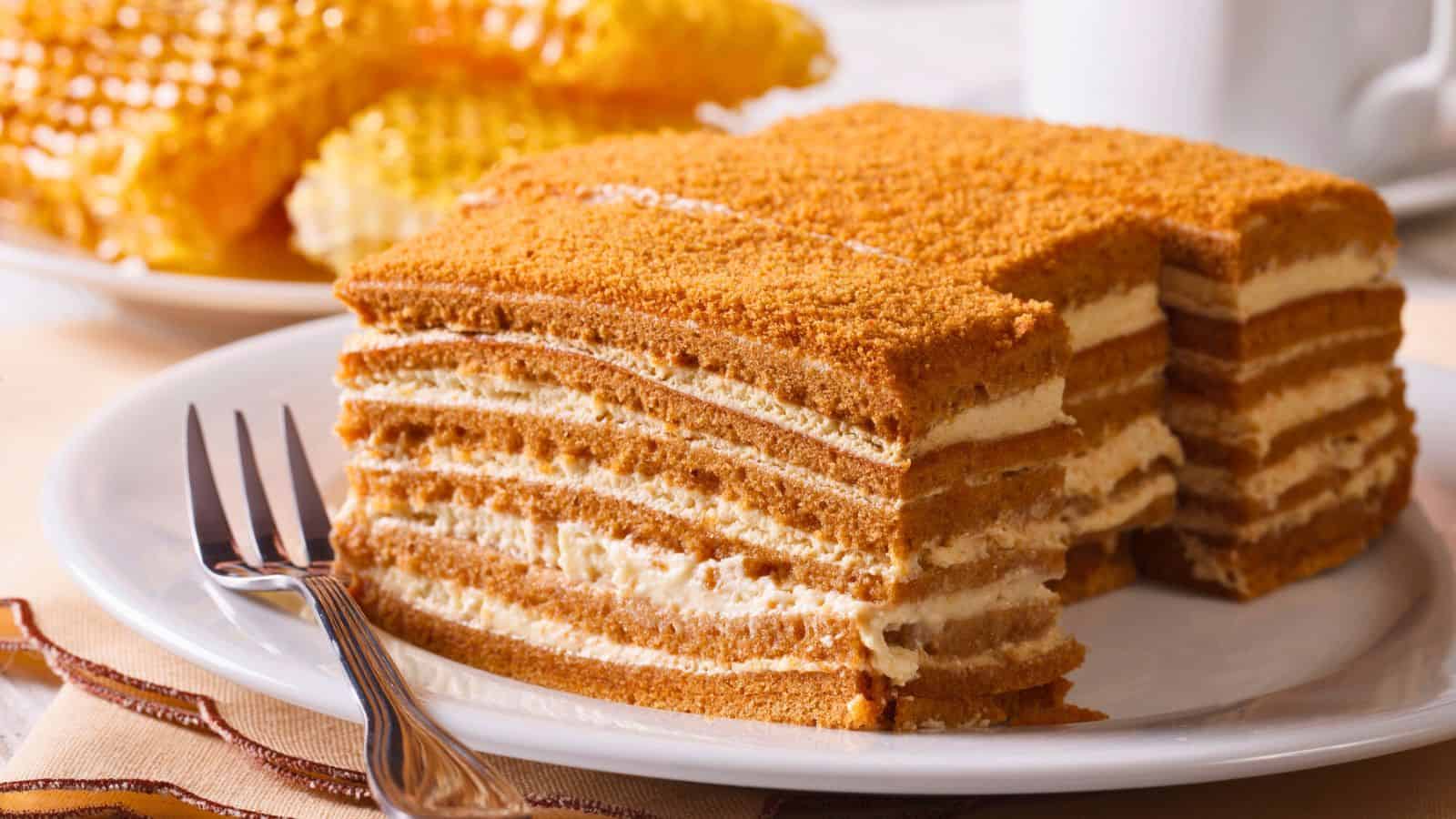 A slice of layered honey cake with cream filling is served on a white plate beside a fork, with pieces of honeycomb and a mug in the background.