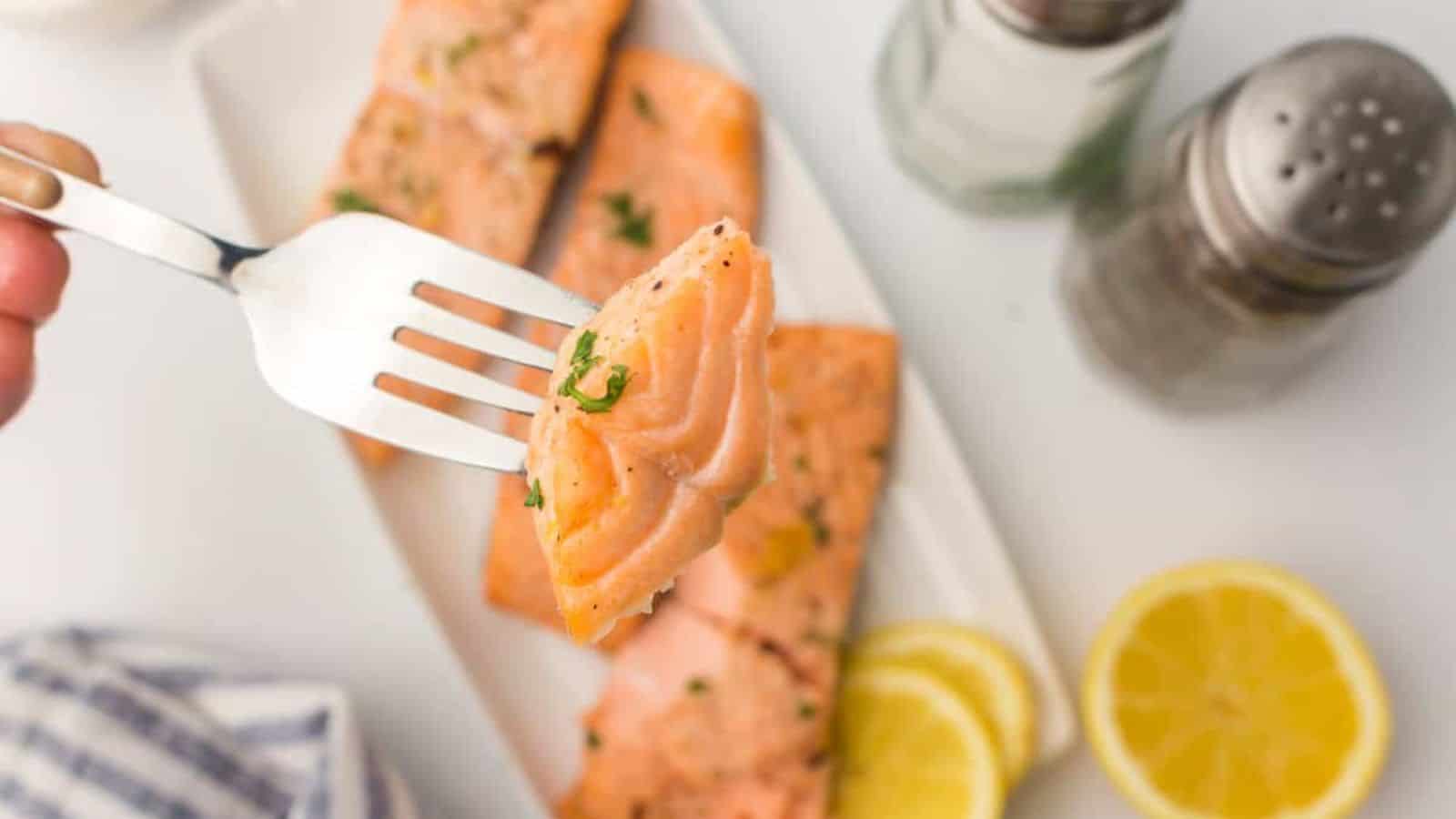 A fork holding a piece of cooked salmon, with more salmon fillets, lemon slices, and seasoning shakers in the background.