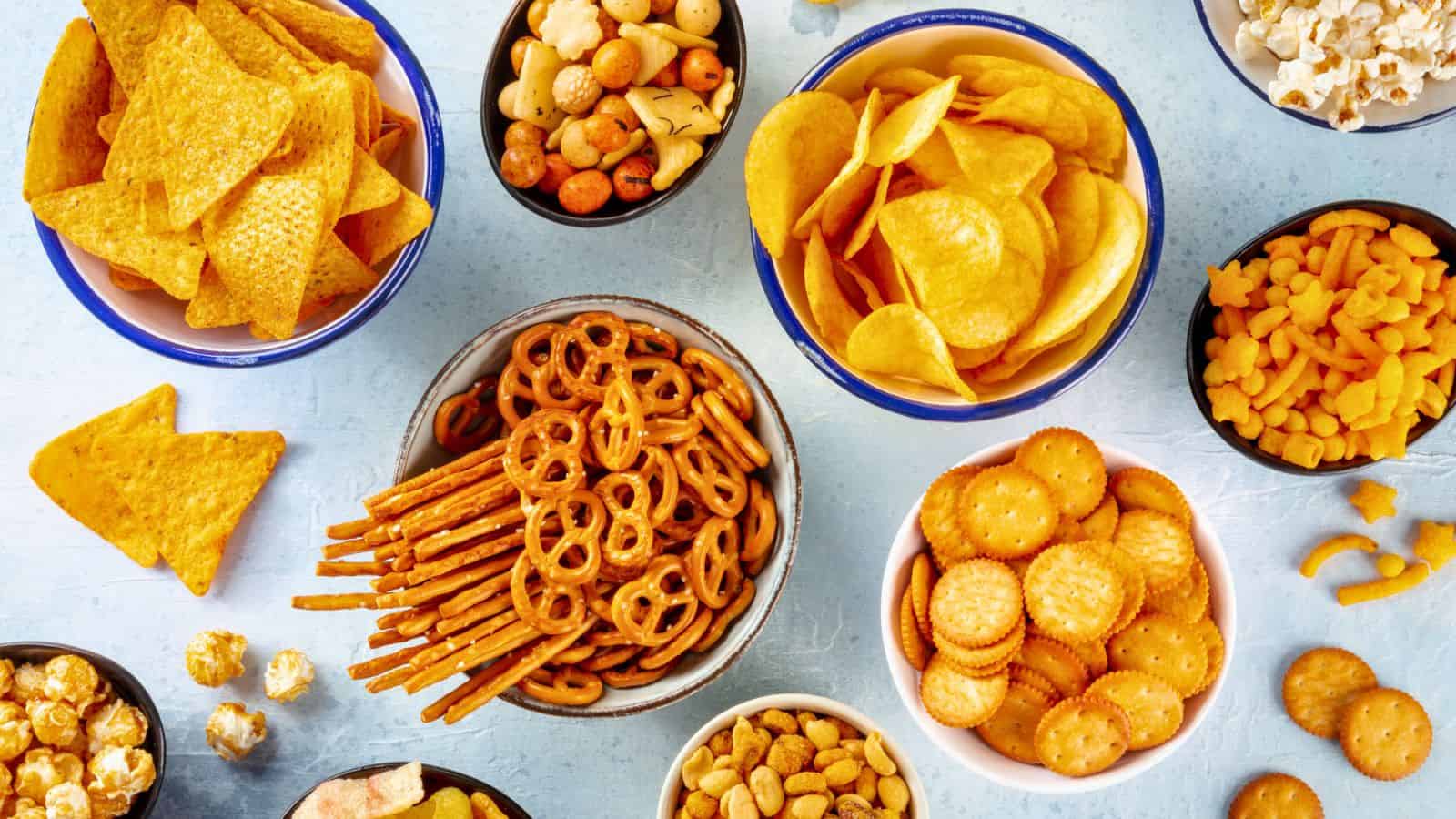 Assorted bowls of snacks are arranged on a light blue surface, including potato chips, tortilla chips, pretzels, cheese crackers, popcorn, mini crackers, and mixed snack nuts.