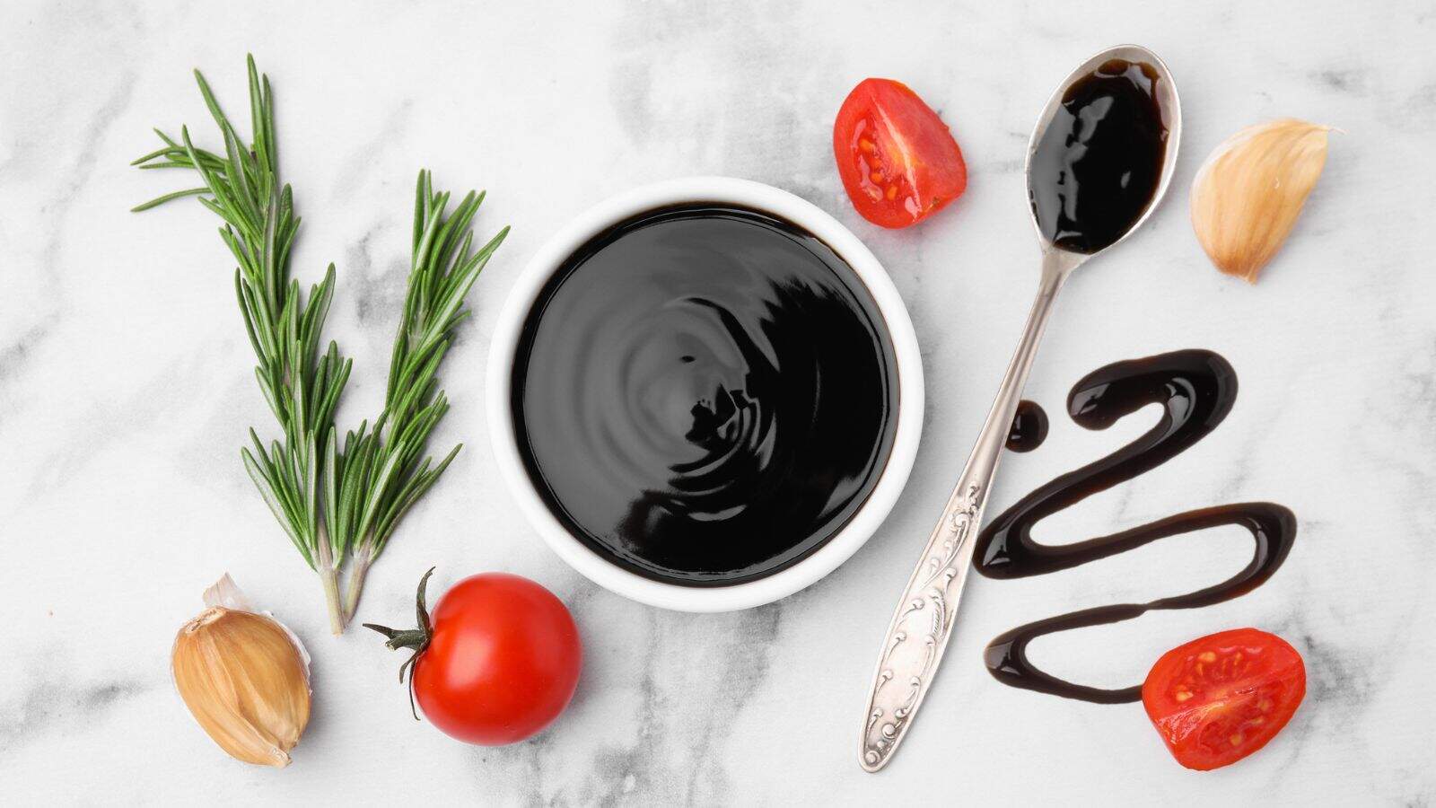 A bowl of thick dark sauce sits on a marble surface, surrounded by a sprig of rosemary, garlic cloves, tomato halves, a whole tomato, and a spoon with sauce drizzled next to it.