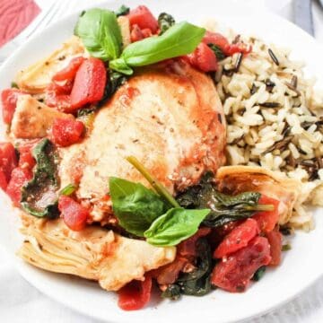 A white plate with a serving of chicken breast topped with tomatoes, spinach, and artichokes, garnished with fresh basil, next to a portion of wild rice. A fork, knife, and red napkin are beside the plate.