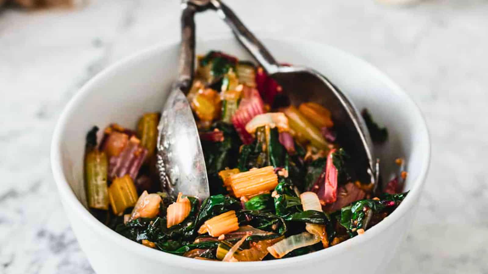 A white bowl filled with cooked Swiss chard stems and leaves sits on a light surface, with metal serving tongs resting inside the bowl.