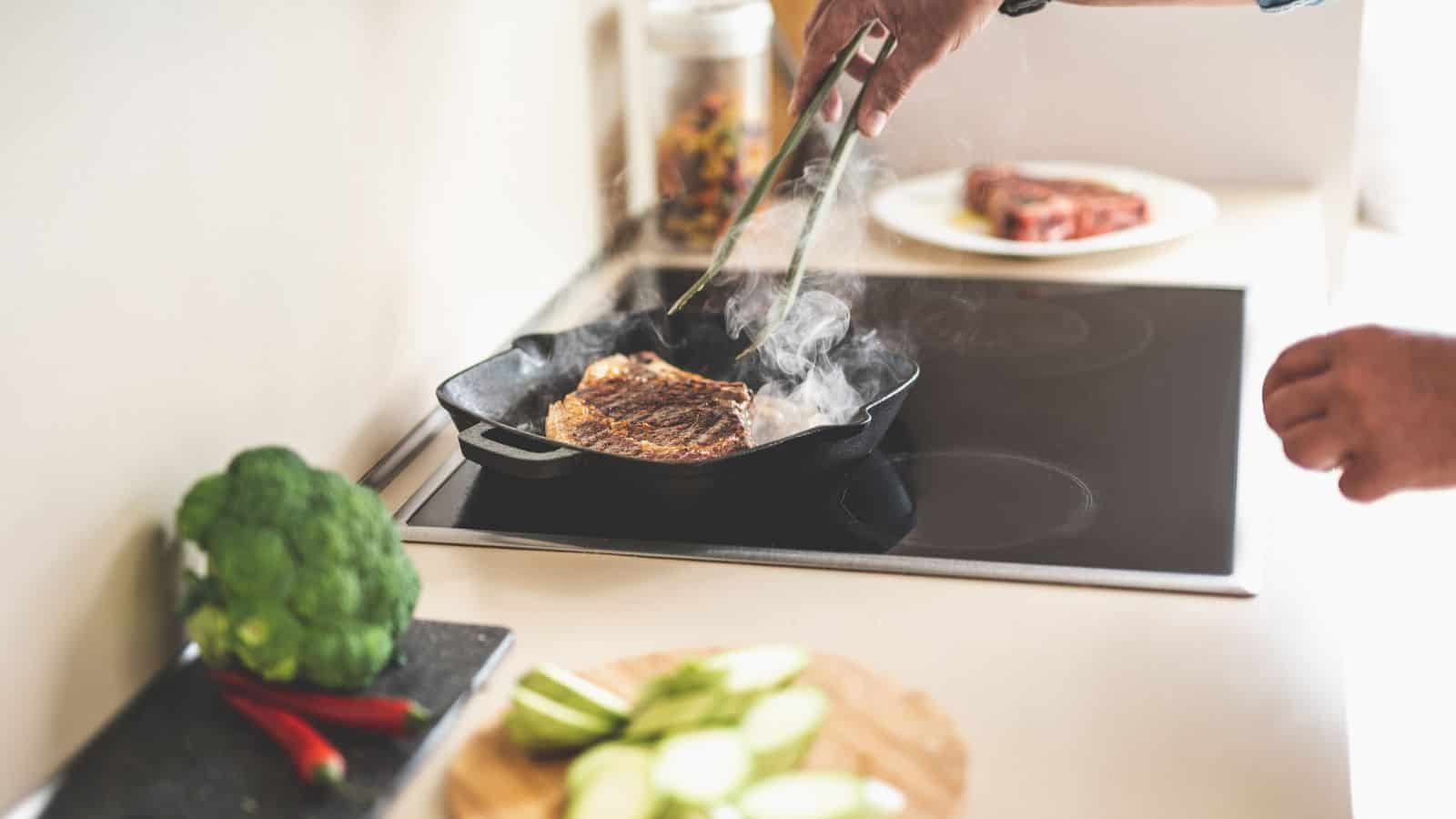 A person uses tongs to cook a steak in a cast iron skillet on a stovetop. Steam or smoke rises from the pan. Vegetables, including broccoli and sliced zucchini, are on the counter nearby.