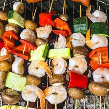 Several skewers of shrimp, red bell pepper, zucchini, and mushrooms are arranged on a grill with other food items in the background—a perfect example of budget cooking that brings big flavors without breaking the bank.