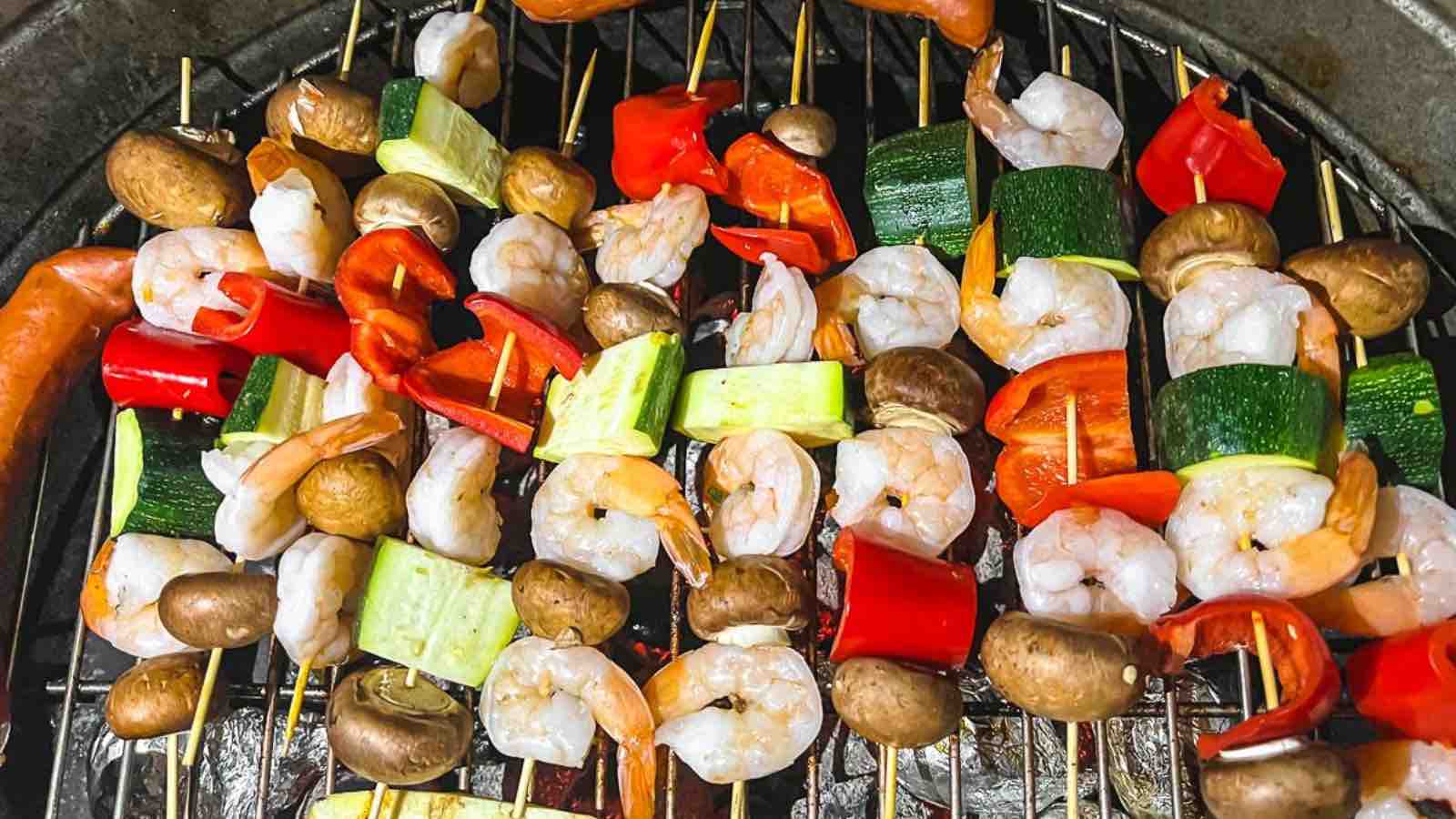 Several skewers of shrimp, red bell pepper, zucchini, and mushrooms are arranged on a grill with other food items in the background—a perfect example of budget cooking that brings big flavors without breaking the bank.