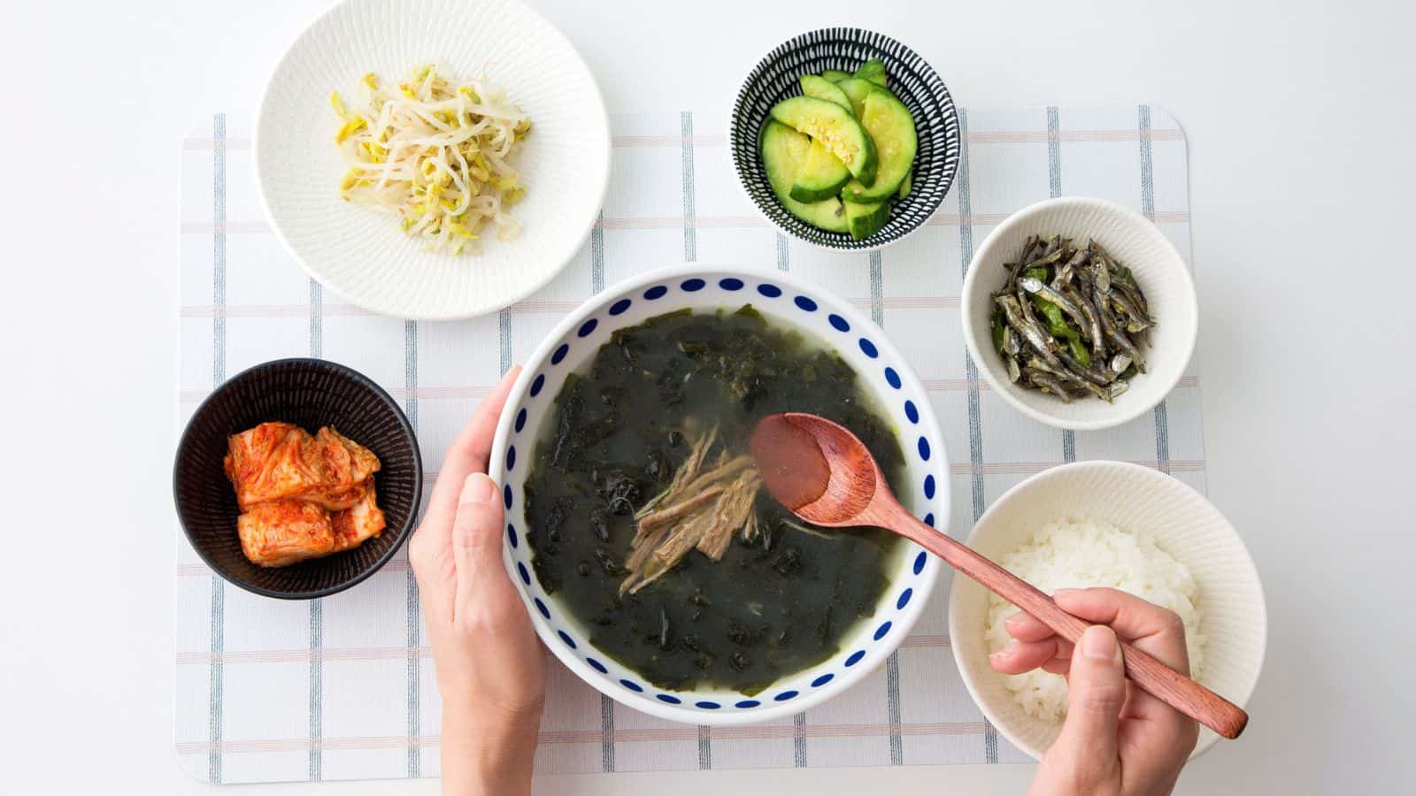 A person holds a bowl of Korean seaweed soup with a wooden spoon, surrounded by small dishes of rice, kimchi, cucumbers, bean sprouts, and seasoned anchovies on a checkered placemat.