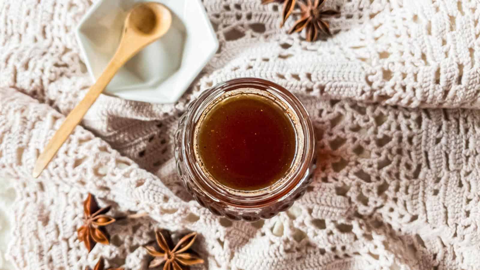 Chai syrup in glass jar on crochet lace fabric with star anise and wooden spoon in white geometric bowl.