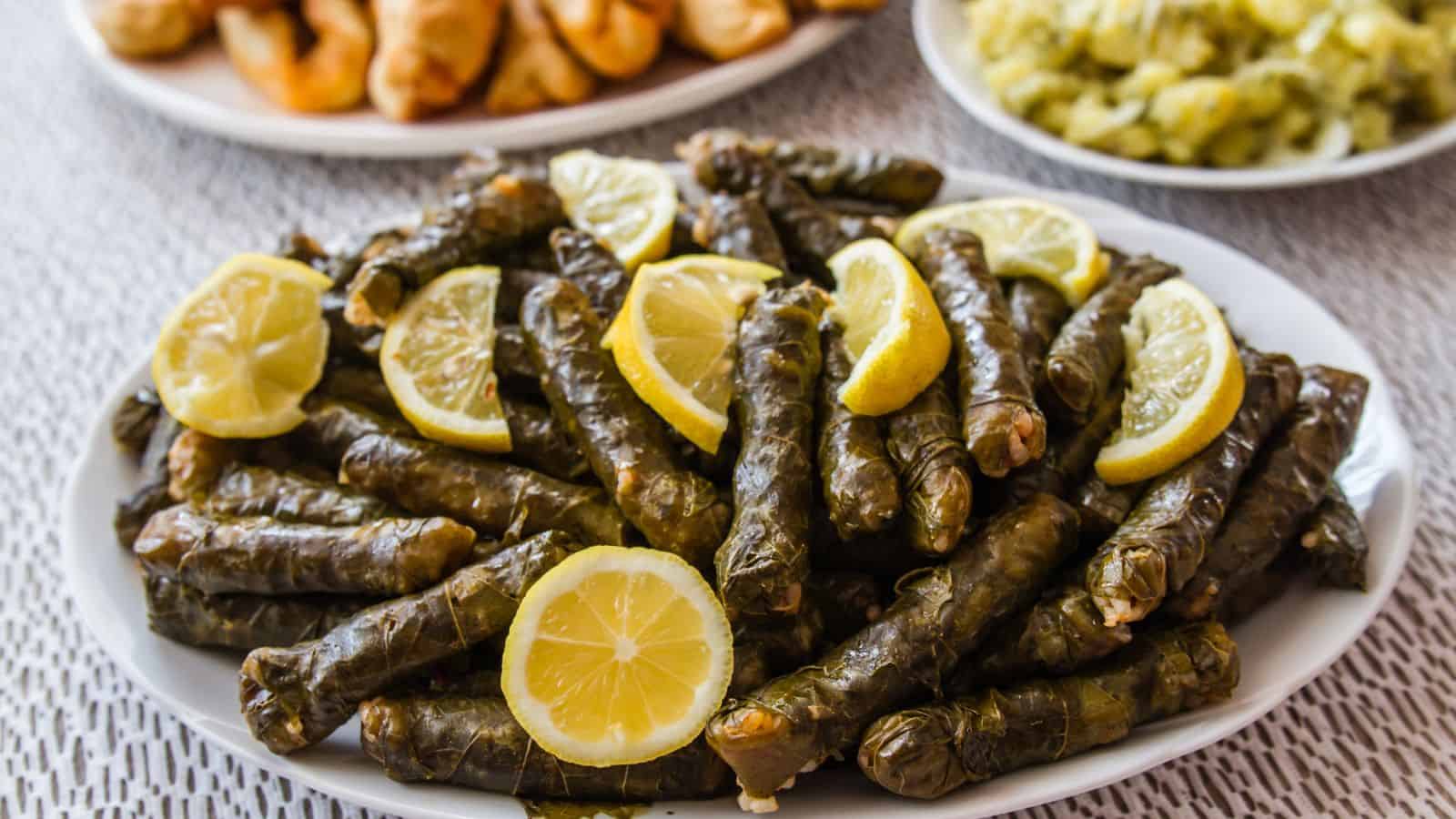 A white plate filled with stuffed grape leaves, each rolled tightly and garnished with slices of lemon. Other plates of food are blurred in the background on a textured tablecloth.