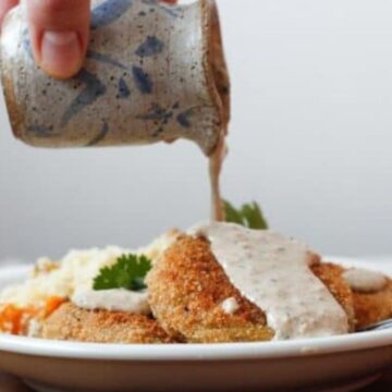 A hand pours creamy sauce from a small ceramic pitcher onto fried green tomatoes on a white plate, garnished with herbs and served with a side of mashed potatoes.