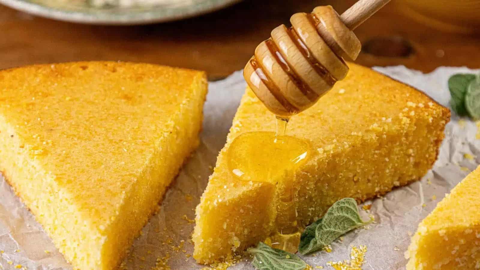 A honey dipper drizzles honey onto two triangular slices of cornbread resting on parchment paper, with a sprig of green herbs nearby.