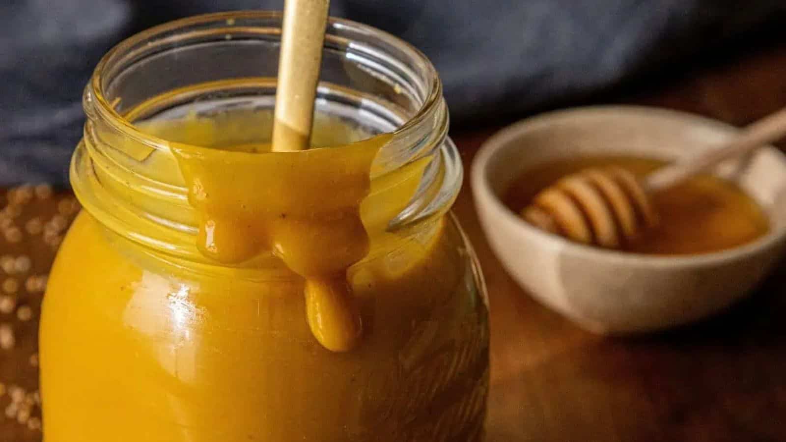 A glass jar filled with thick, golden yellow Carolina Mustard BBQ Sauce, some dripping down the side; a spoon rests inside. In the background, there's a small bowl of honey with a honey dipper—perfect for drizzling over pulled pork.