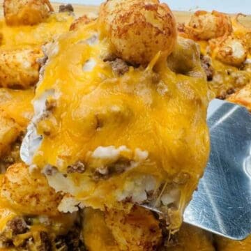 A metal spatula lifts a serving of a casserole with melted cheddar cheese, ground beef, tater tots, and mashed potatoes from a glass baking dish.