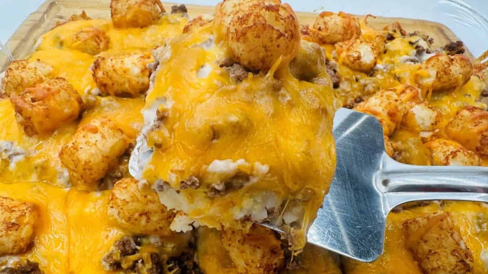 A spatula lifts a portion of a baked casserole featuring tater tots, ground beef, and melted cheddar cheese from a glass dish.